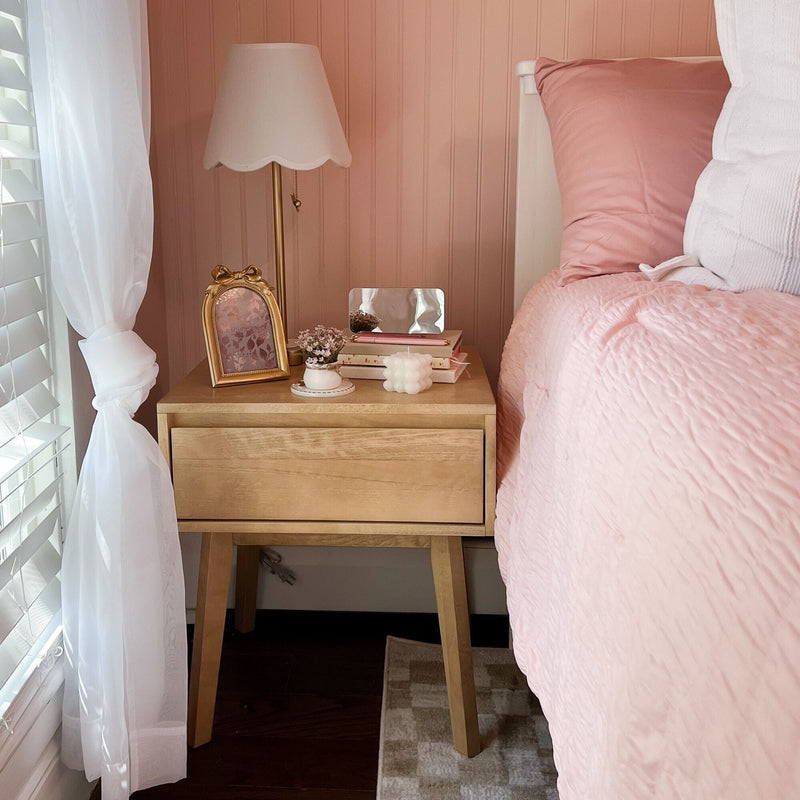 Cozy pink bedroom featuring a wooden mid-century bedside table with a scalloped white lamp, gold frame, stacked books, small vase of flowers and blush quilted bedding