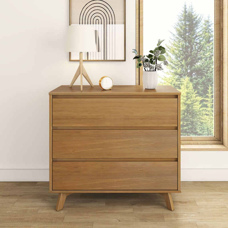 Mid-century modern three-drawer oak dresser in a minimalist bedroom, natural wood finish styled with a tripod table lamp, gold alarm clock, white ceramic vase with greenery and large window forest view