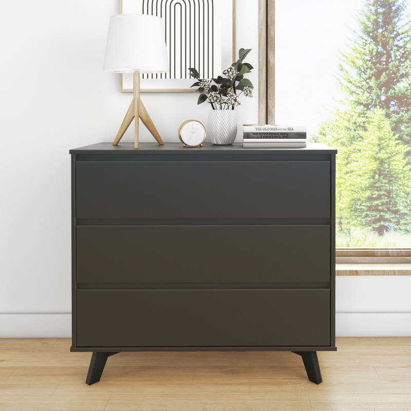 Modern black three-drawer dresser with mid-century legs styled with a wooden lamp, clock, white vase, books and abstract art in a bright minimalist bedroom by a window overlooking trees