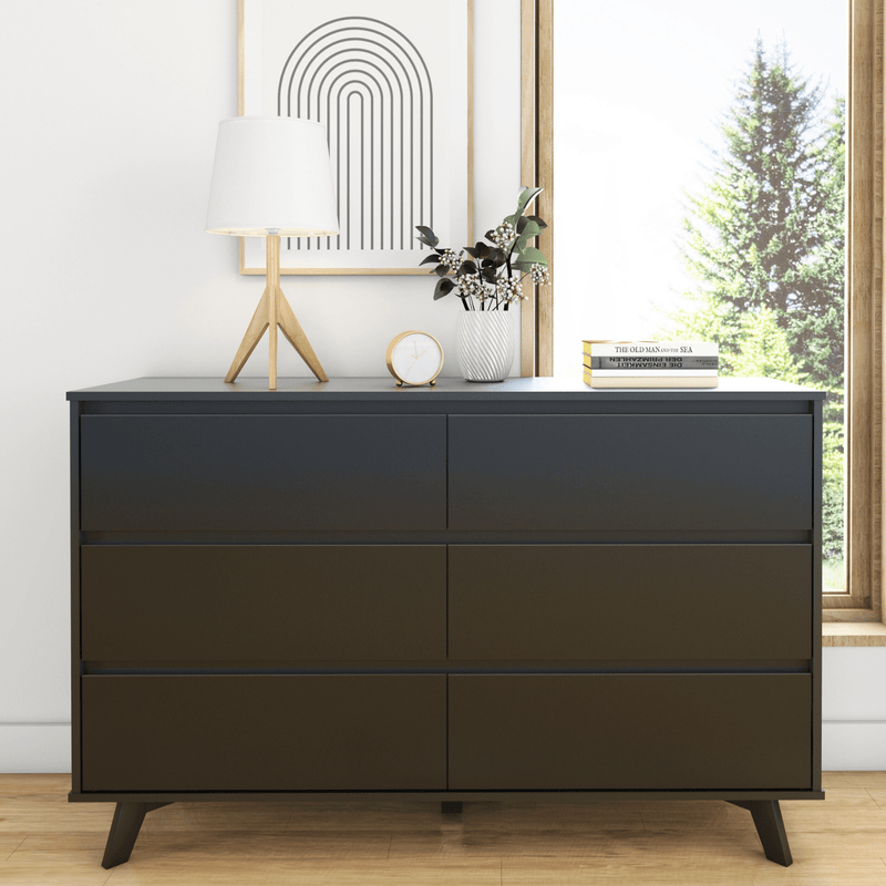 Modern black six-drawer dresser with mid-century legs, styled with a wooden tripod lamp, potted plant, clock and stacked books by a sunny window, minimalist bedroom decor