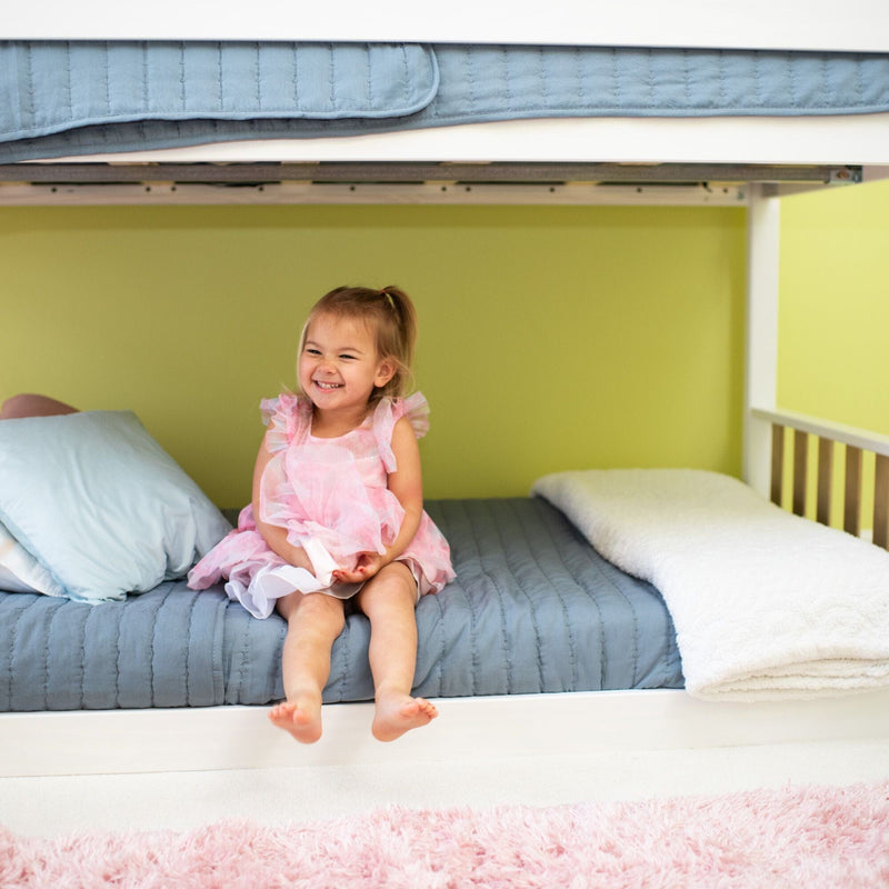Smiling toddler girl in a pink dress sitting on a blue-quilted lower bunk bed in a green nursery with pillows and a pink shag rug — kids bedroom decor and bedding