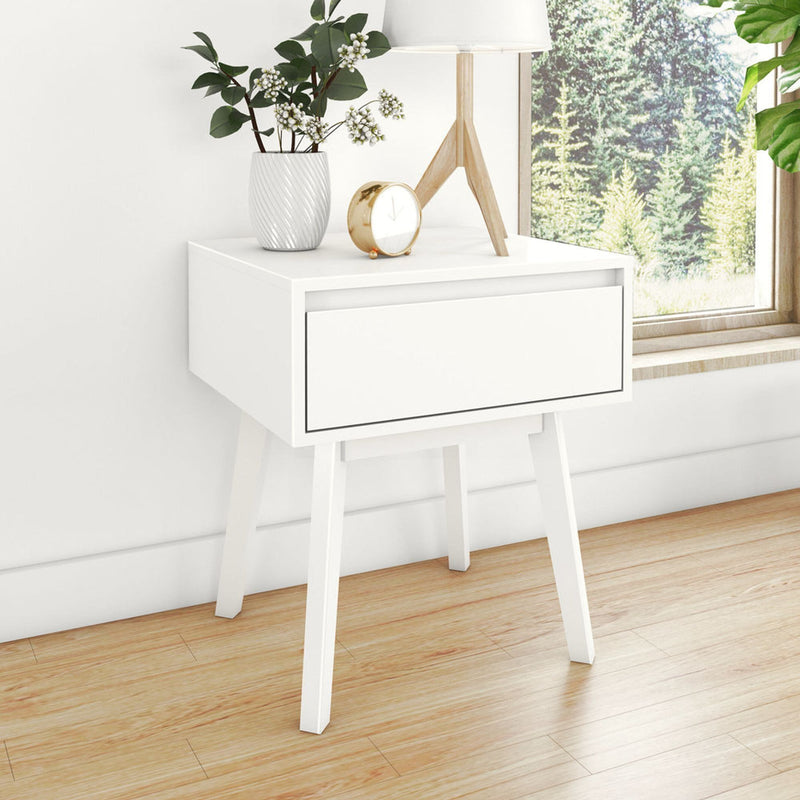 White modern bedside table nightstand with single drawer and wooden legs, lamp, vase with greenery and gold alarm clock on hardwood floor by window with forest view
