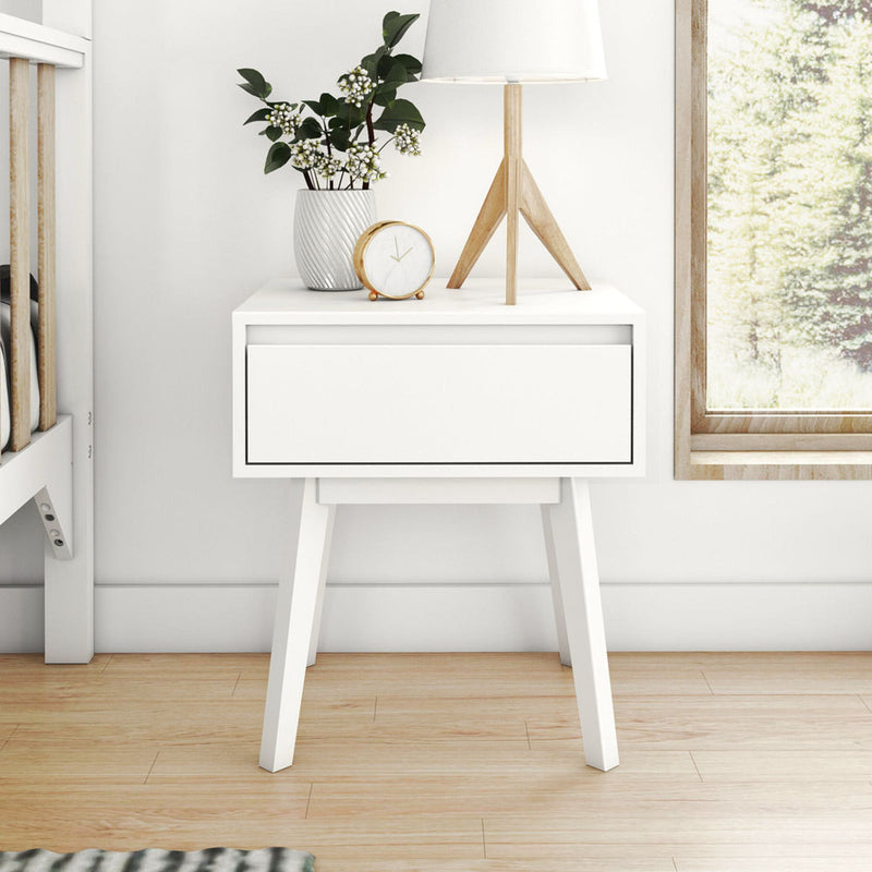 White mid-century modern nightstand with single drawer, wooden tripod lamp, potted green plant and gold-accent alarm clock on hardwood floor by window, minimalist Scandinavian bedroom decor