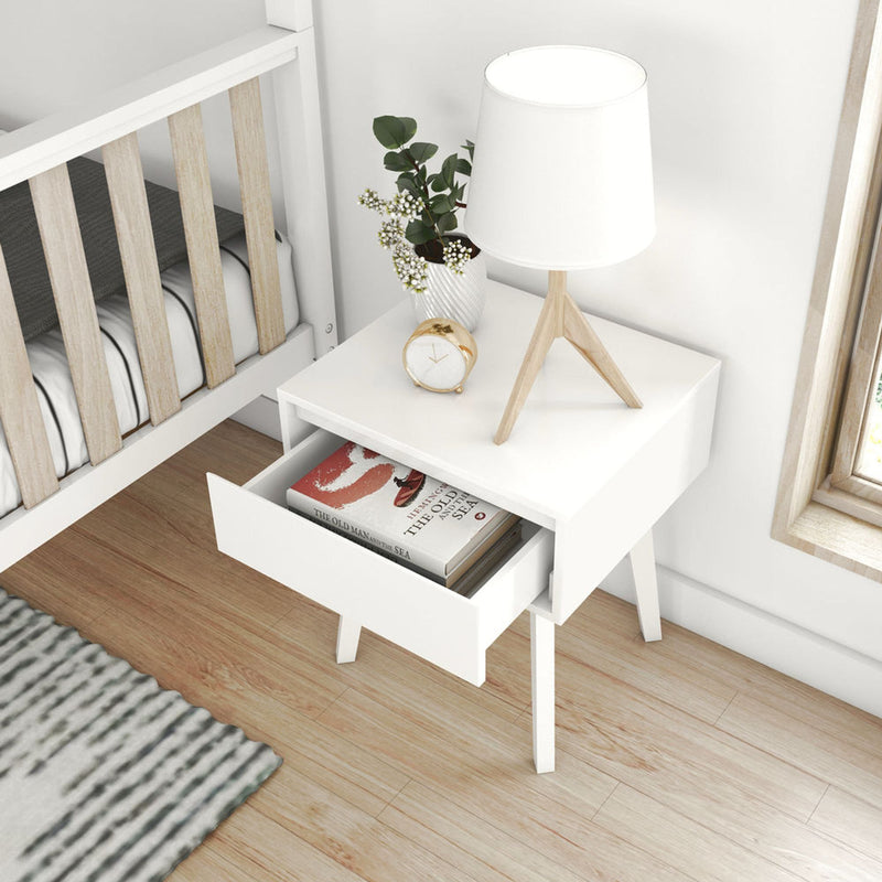 White Scandinavian bedside table with open drawer, wooden tripod lamp, gold alarm clock, vase and book beside baby crib on hardwood floor