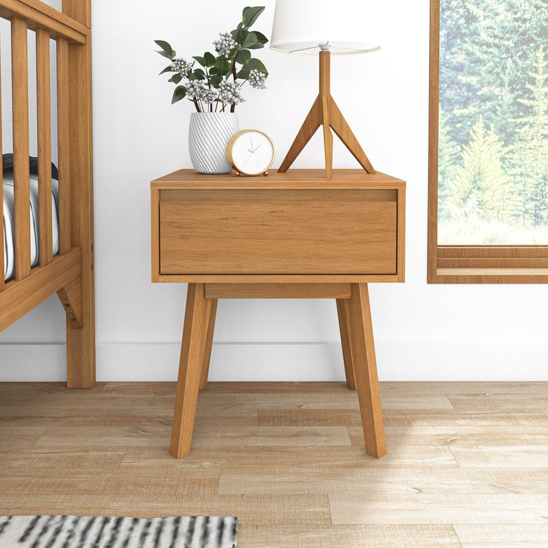 Mid-century modern wooden nightstand in nursery with lamp, gold alarm clock, white vase, crib and hardwood floor by window with forest view