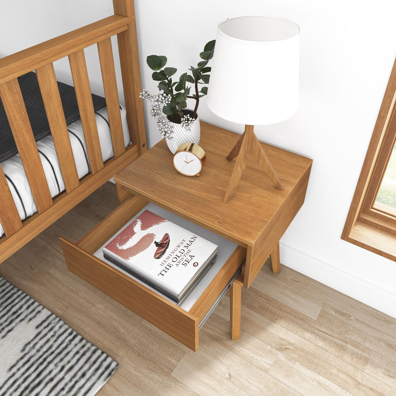 Mid-century wooden nightstand with open drawer holding "The Old Man and the Sea", white tripod table lamp, potted plant and gold alarm clock on light hardwood bedroom floor