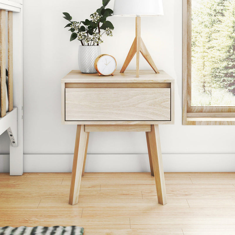 Light wood Scandinavian bedside table with single drawer, tripod table lamp, small potted plant and gold alarm clock by a window in a minimalist bedroom.