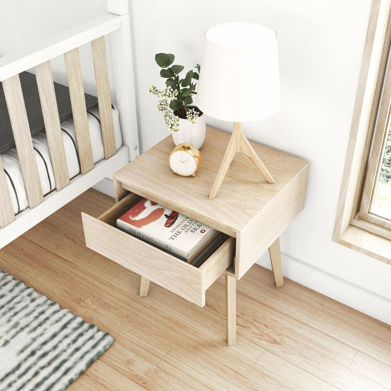 Light wood Scandinavian bedside table with white tripod lamp, gold alarm clock, potted greenery and open drawer of books next to a white slatted crib on hardwood floor, minimalist nursery bedroom decor