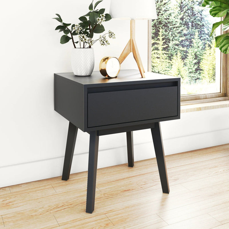 Matte black mid-century modern bedside nightstand with single drawer and tapered angled legs, styled with white vase, brass clock and wooden tripod lamp on light hardwood floor by a window