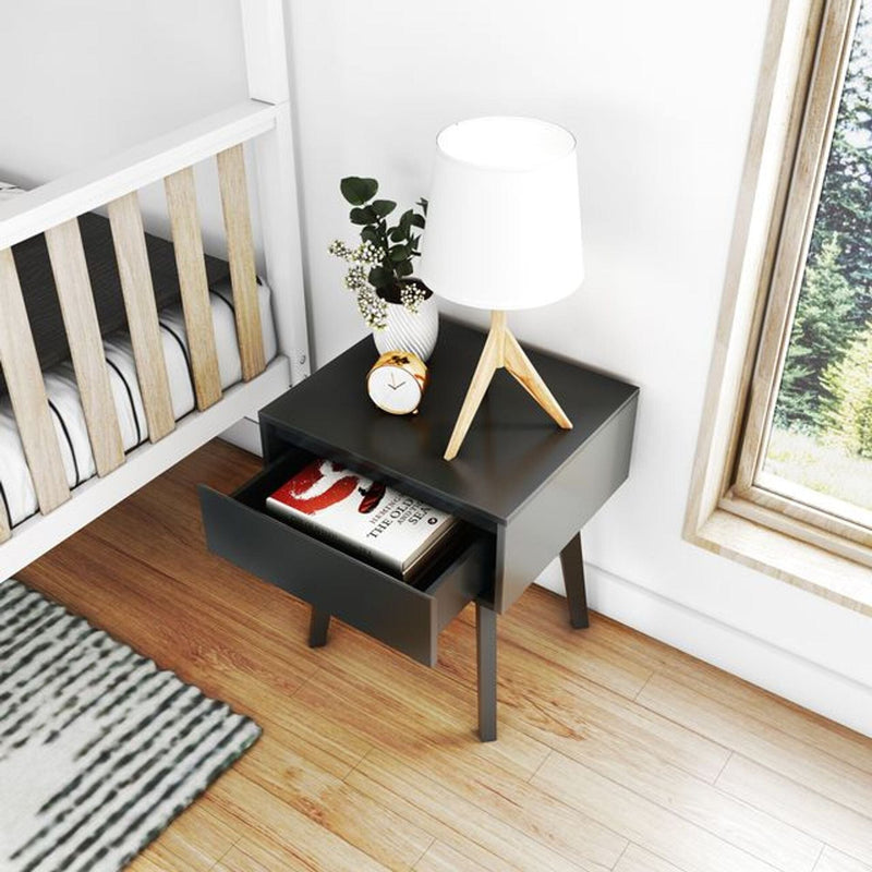 Modern black bedside nightstand with open drawer holding books, wooden tripod lamp, alarm clock and potted plant beside white crib on hardwood floor by window