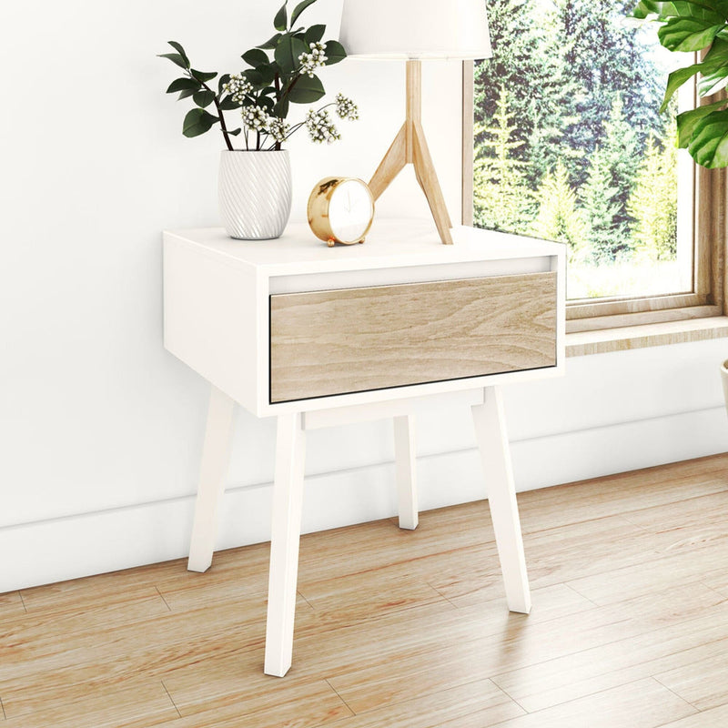 Mid-century modern white nightstand with natural wood drawer, table lamp, potted plant and gold alarm clock on hardwood floor by window showing forest view