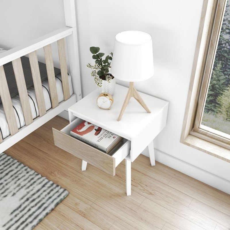 White Scandinavian nursery nightstand with tripod table lamp, gold alarm clock, potted greenery and open drawer beside wooden crib