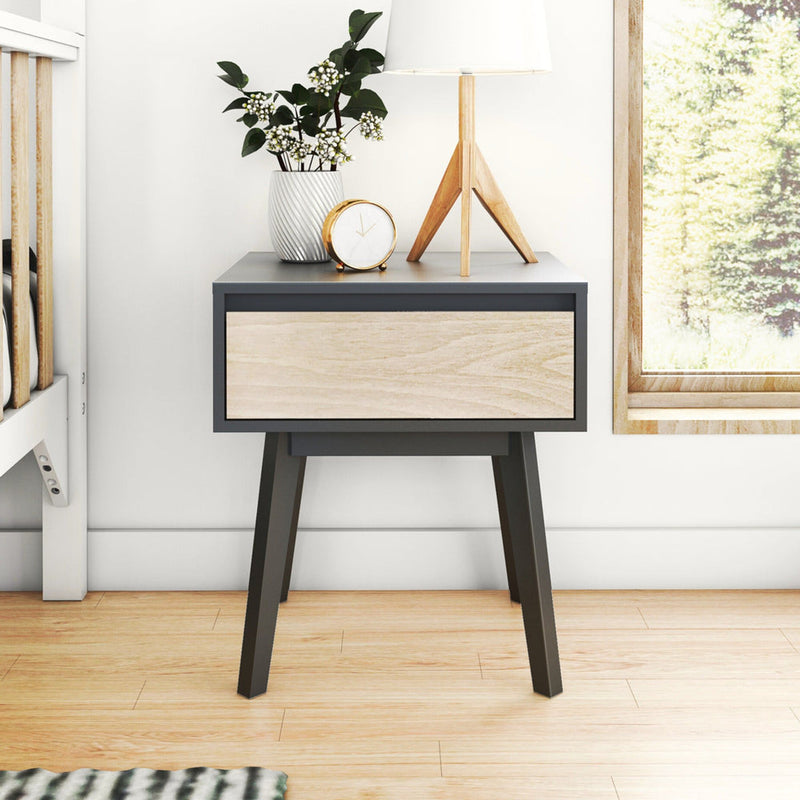 Modern mid-century bedside nightstand in gray and light wood with drawer, tripod lamp, gold alarm clock, potted plant and hardwood floor in bright bedroom by window