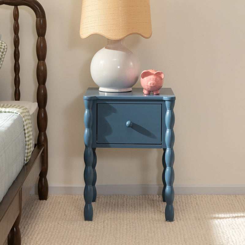 Blue wooden bedside table nightstand with turned legs and drawer, white ceramic lamp with beige shade and pink piggy bank beside wooden spindle bed in cozy bedroom