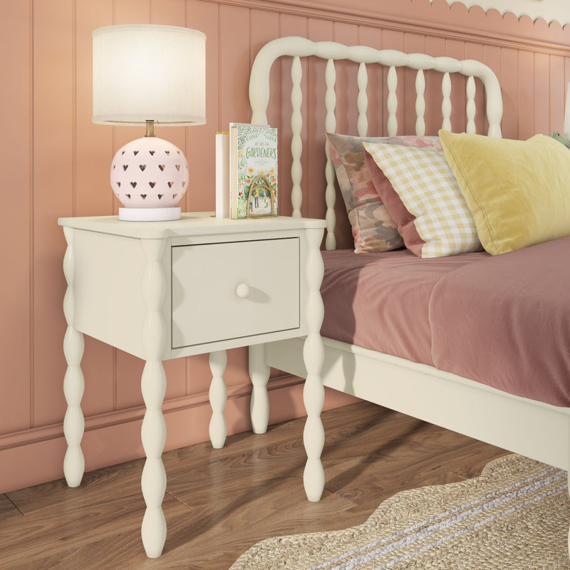 White spindle-leg nightstand with drawer and pink heart-pattern lamp beside cream spindle bed with mauve bedding and pastel pillows against a pink paneled wall on wood floor with woven rug, cozy children's bedroom