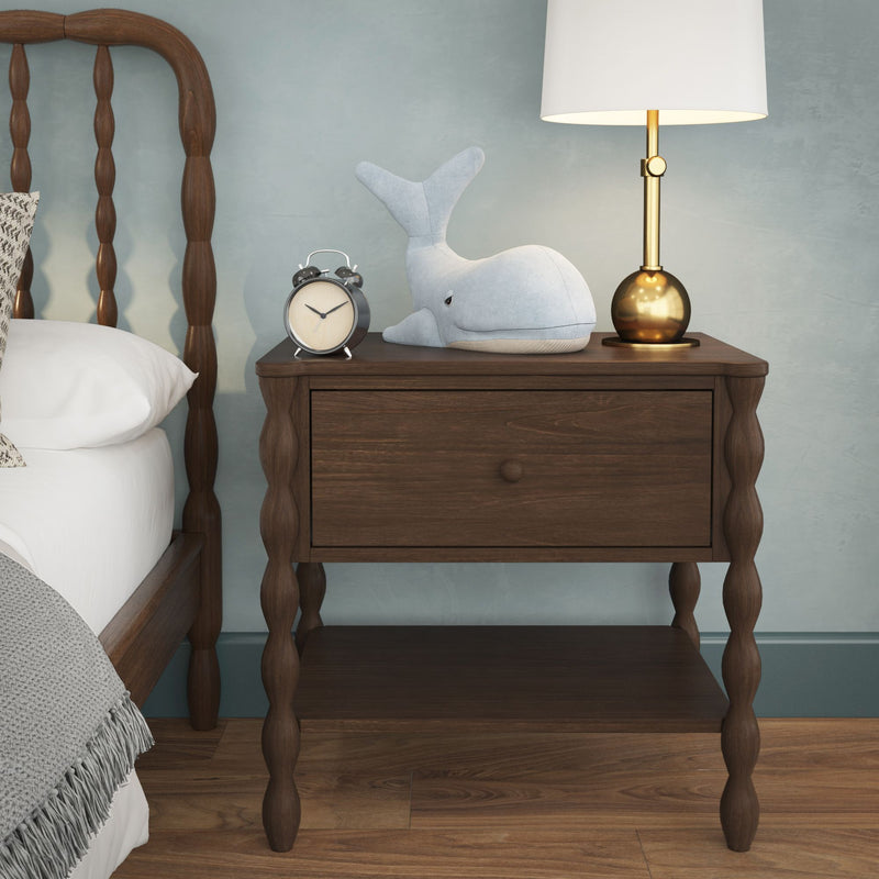Dark wood nightstand with drawer topped by brass lamp, vintage alarm clock and blue whale plush beside spindle bed in modern bedroom