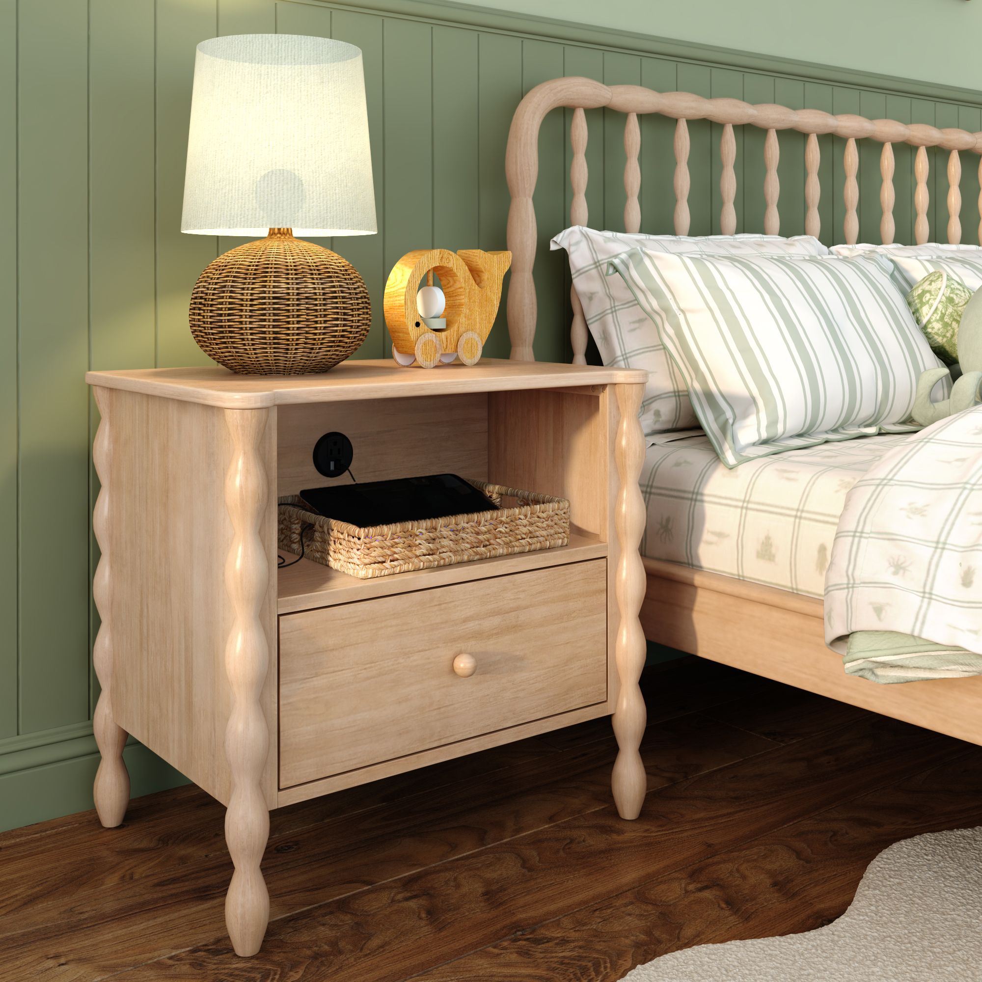 A light-wood finish Jenny Lind style nightstand next to a matching bed in a green room. The nightstand has a shelf with a charger port, holding a basket containing a device. On top of the nightstand is a lamp and decor.