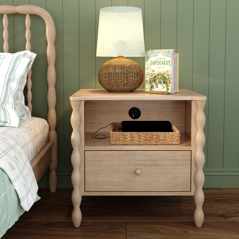 Light wood bedside table with turned legs, woven rattan lamp, children's books and wicker charging tray beside a bed against a green paneled wall