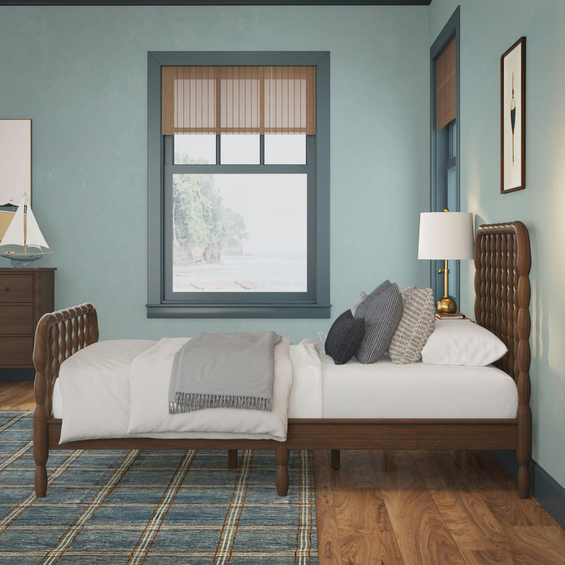 Coastal bedroom interior with wooden spindle bed and tufted headboard, white layered bedding and gray throw, patterned pillows, blue plaid rug, wooden nightstand with sailboat decor and brass lamp, teal walls and bamboo blinds framing an ocean view.