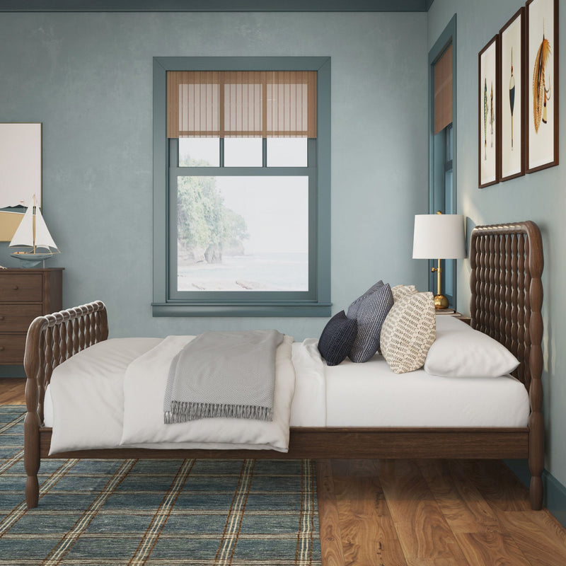 Coastal bedroom interior with wooden spindle bed and tufted headboard, blue-gray walls, window framing a beach view, nautical decor, plaid area rug and bedside lamp