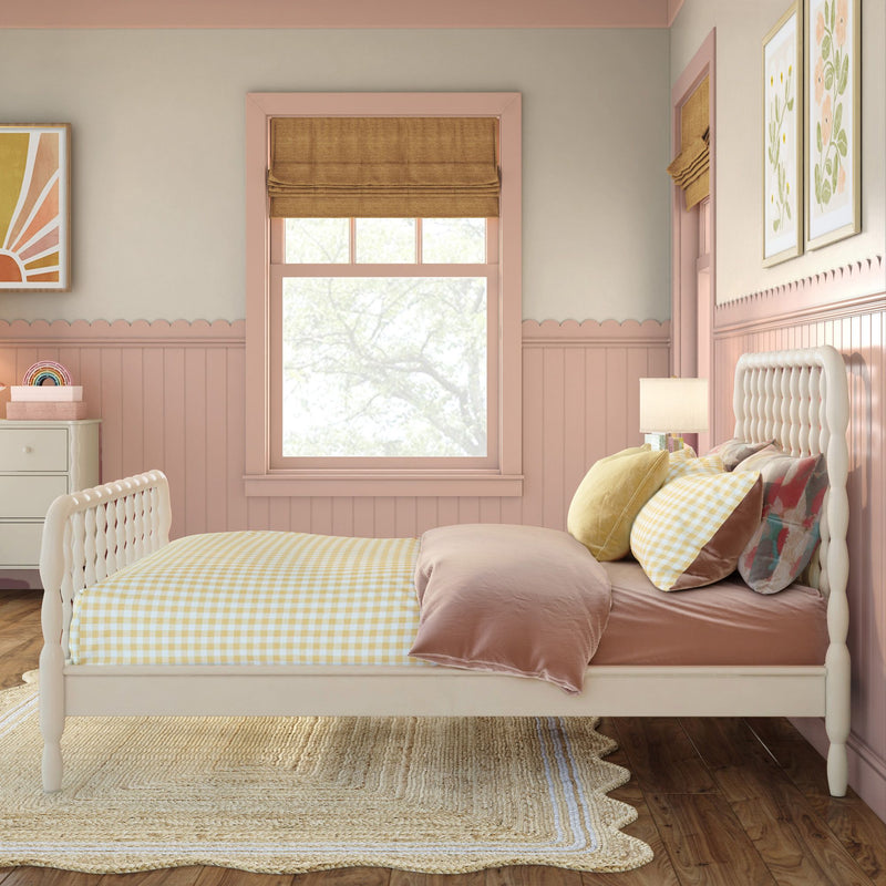 Pastel pink children's bedroom with tufted white headboard, yellow gingham bedding, layered throw pillows, pink wainscoting, Roman shade window, framed floral art and bedside lamp