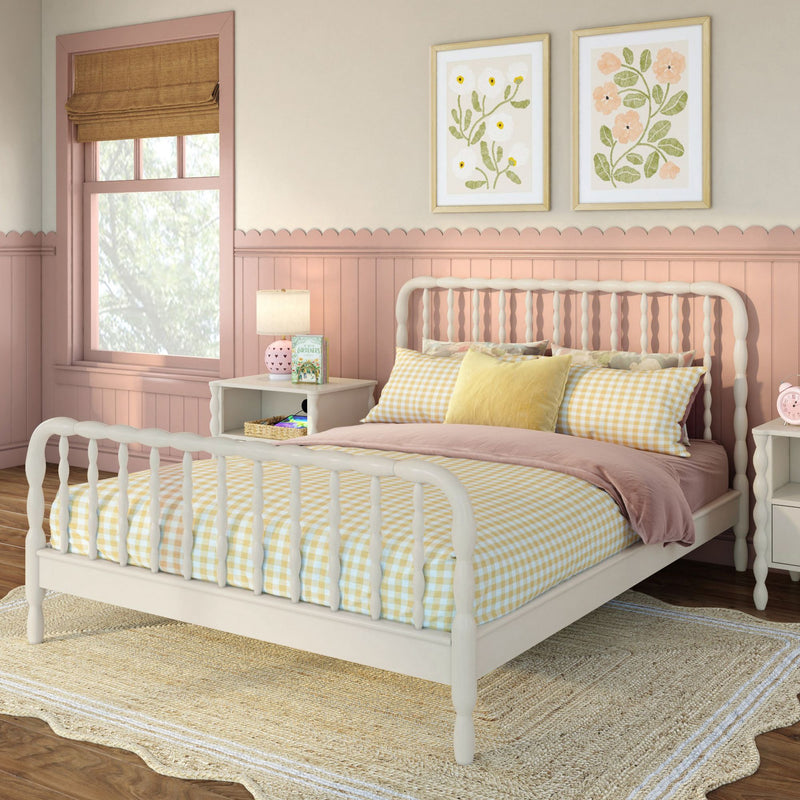 Pastel pink nursery with white spindle bed, yellow gingham bedding, scalloped wainscoting, framed floral prints and bedside lamp