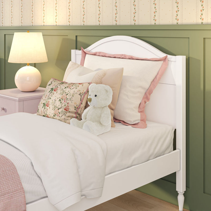 Cozy girl's bedroom with white wooden bed frame, pastel pink and floral pillows, plush teddy bear, round bedside lamp and green wainscoting