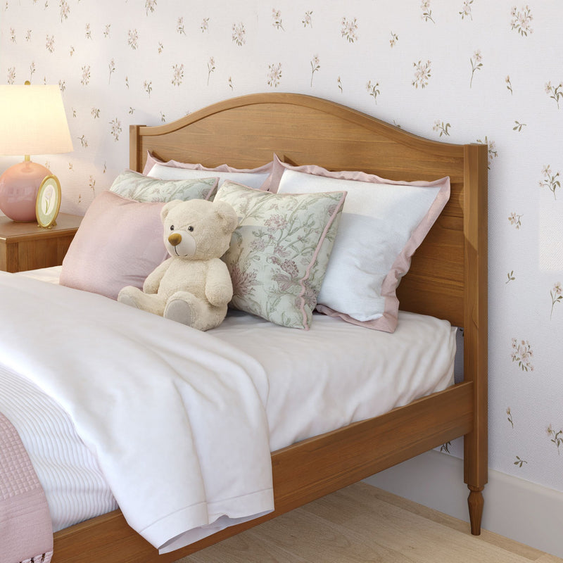 Cozy children's bedroom with wooden headboard, pink floral pillows, plush teddy bear, white duvet and bedside lamp against pastel floral wallpaper