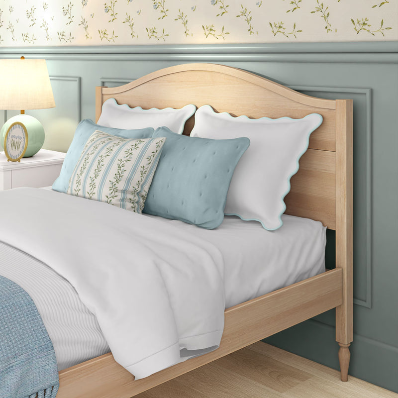 Cozy coastal cottage bedroom interior with light wood headboard, scalloped white pillows with aqua trim, blue throw pillows, floral accent pillow, mint lamp on white nightstand and pastel wainscoting