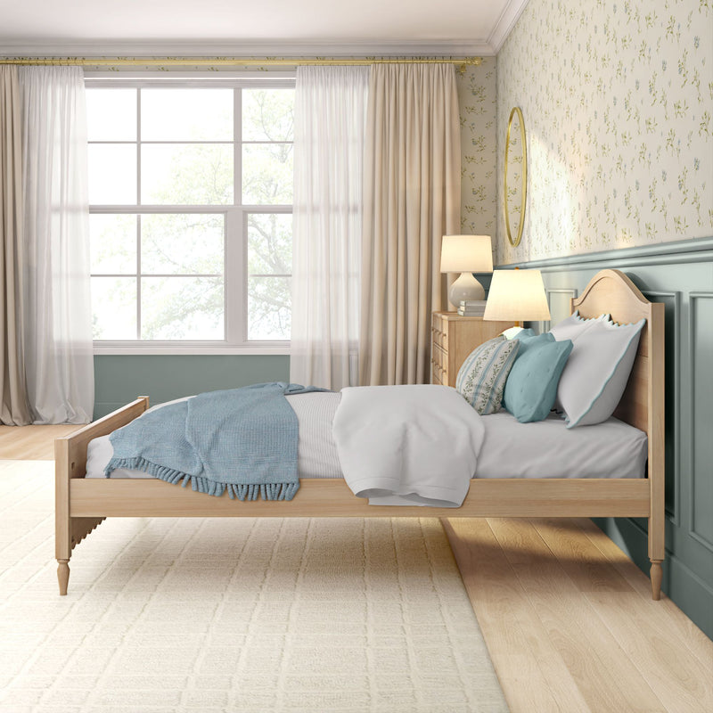 Light-filled cozy bedroom with wooden bed, blue and white bedding and throw, floral wallpaper, large window with sheer beige curtains and bedside lamp