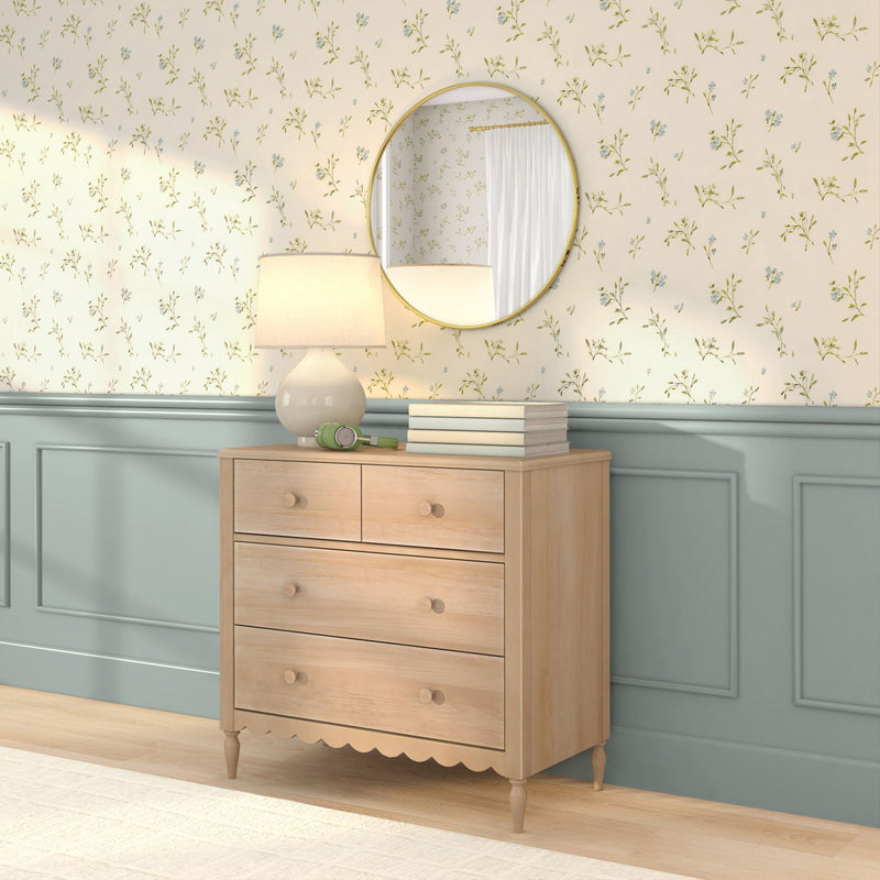 Light wood bedroom dresser with round gold mirror, ceramic table lamp, green headphones and books against floral wallpaper and mint wainscoting