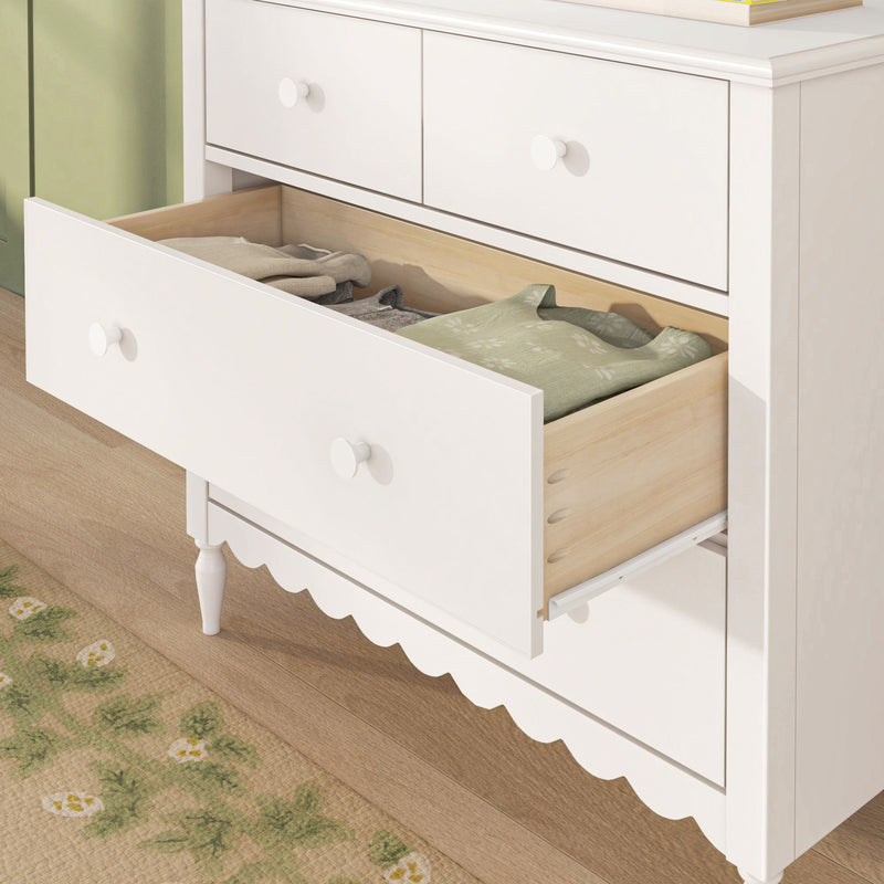 White wooden dresser with open drawer showing folded clothes, round knobs and scalloped base — bedroom or nursery storage furniture