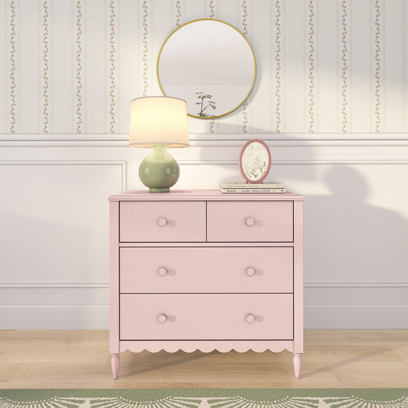 Pastel pink dresser topped with a green ceramic table lamp, gold round mirror and oval frame against floral striped wallpaper and white wainscoting on light wood floor