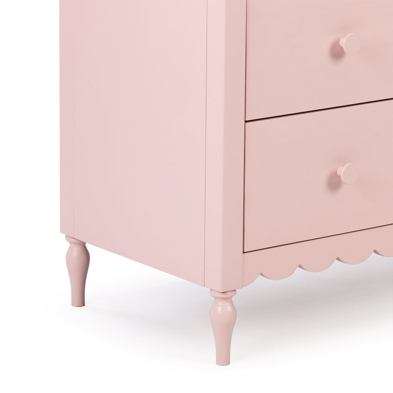 Close-up of a pink painted wooden dresser corner showing drawers with round knobs, scalloped skirt and turned spindle legs