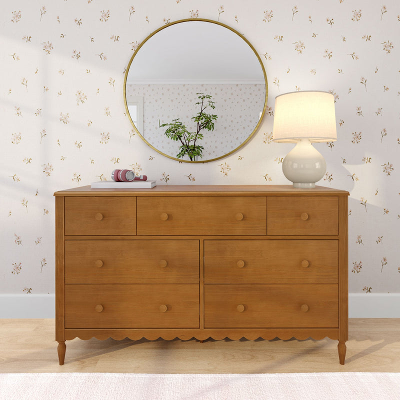 Wooden dresser with round gold wall mirror reflecting a potted plant, cream table lamp and pink headphones on floral wallpaper, styled bedroom dresser decor