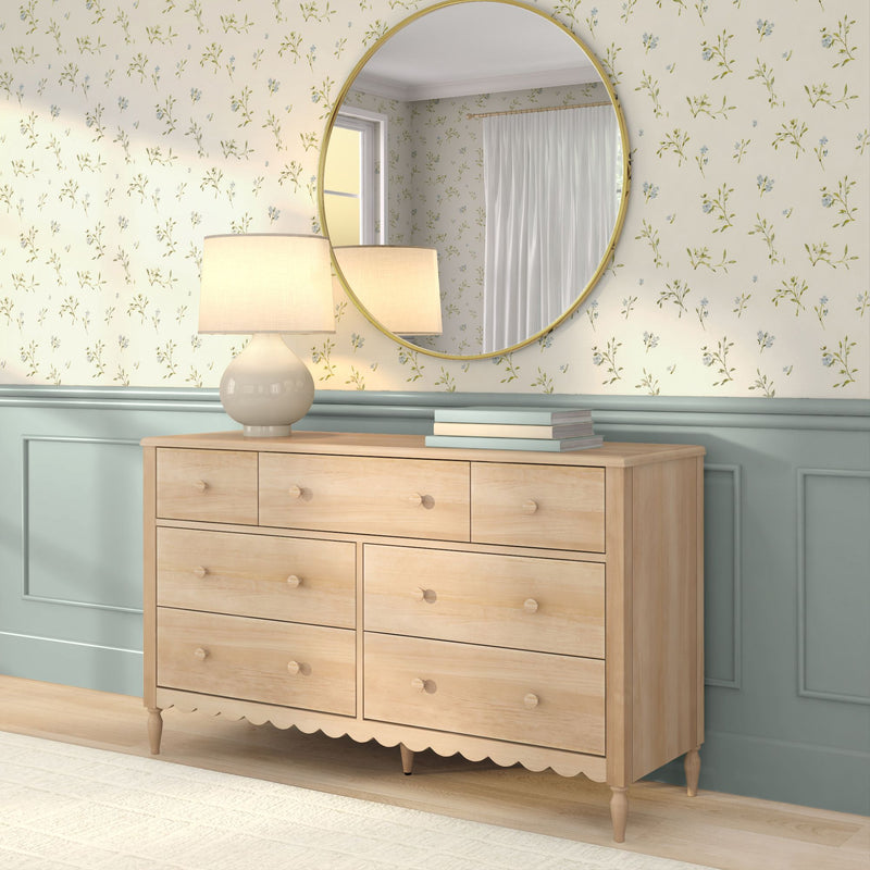 Light wood dresser with round gold mirror, ceramic table lamp and stacked books against floral wallpaper and mint wainscoting, cozy bedroom decor