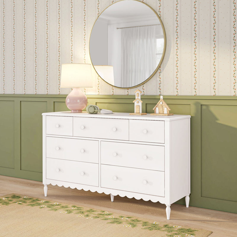 White wooden dresser with round gold mirror, pink lamp, wooden toy houses and headphones against green wainscoting and floral wallpaper, cozy nursery decor