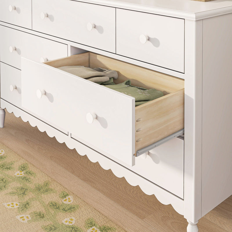 White wooden nursery dresser with open drawer revealing folded baby clothes, round knobs and scalloped skirt on hardwood floor