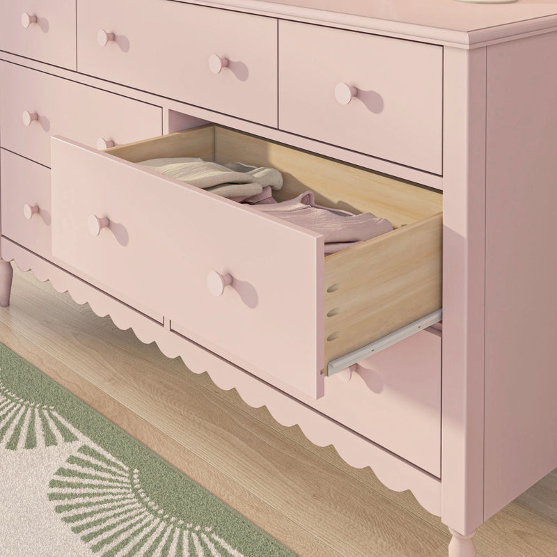 Pink wooden dresser with open drawer revealing folded baby clothes, scalloped trim and round knobs on hardwood floor next to a green patterned rug — nursery storage and children's dresser