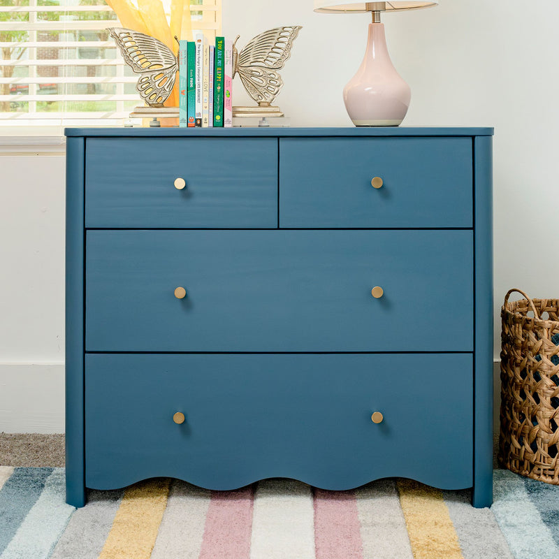 Blue dresser with brass knobs, decorative butterfly bookends holding children's books, pink ceramic table lamp and woven basket on a pastel striped rug in a bright nursery