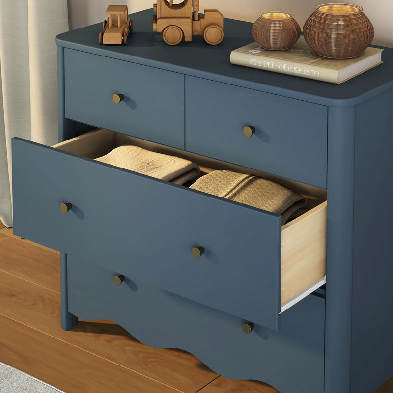 Navy blue wooden dresser with brass knobs and open drawer revealing folded blankets, styled on top with wooden toy cars, an "Art Deco" book and wicker candle holders