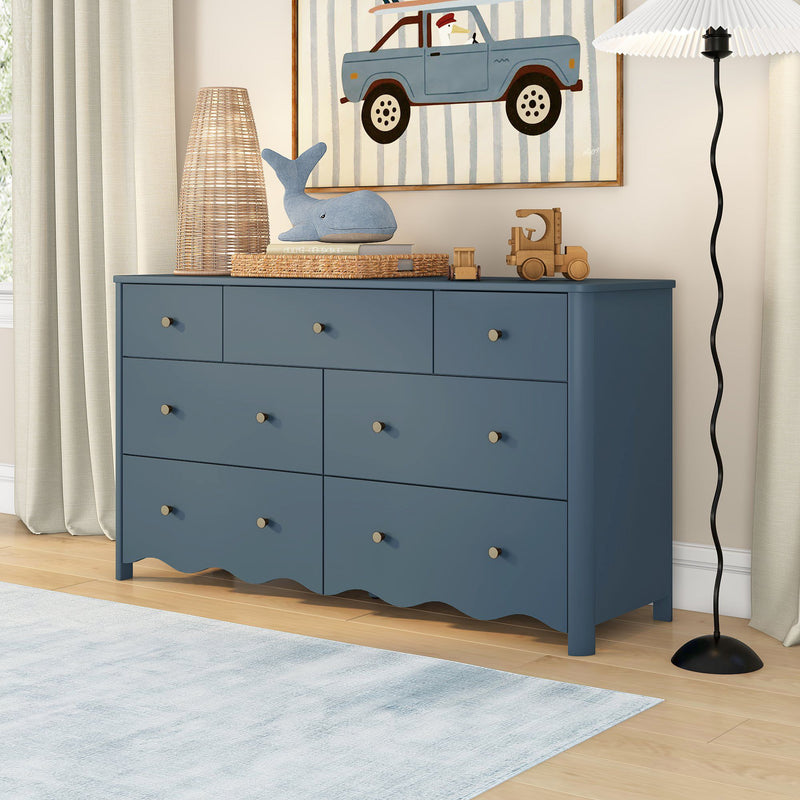 Blue wooden nursery dresser for kids room storage styled with whale plush, wicker lamp, wooden toy truck, framed blue car artwork, neutral curtains and modern floor lamp on hardwood floor