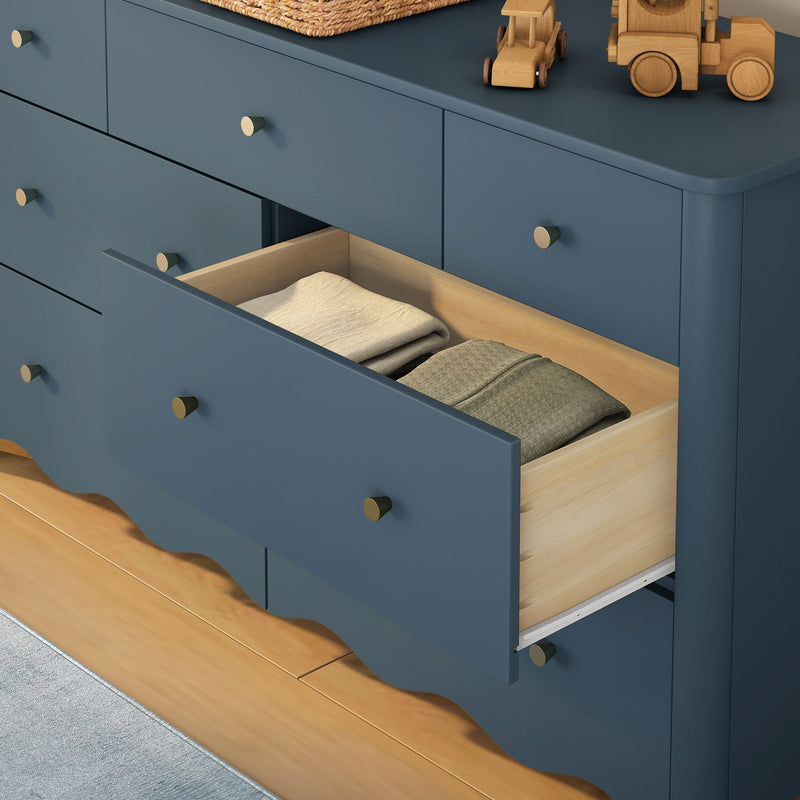 Matte blue modern dresser with open drawer revealing neatly folded clothes, brass knobs, wooden toy trucks and woven basket on top — nursery storage furniture on hardwood floor