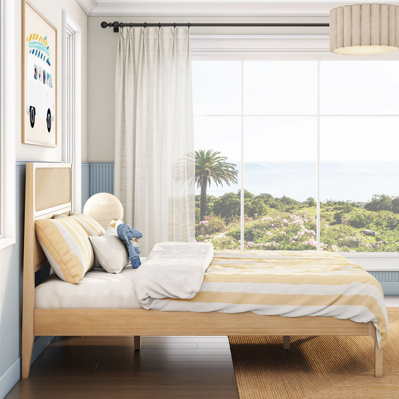 Coastal bedroom with large window and ocean view, natural wood bed with yellow-striped bedding, sheer curtains, palm tree outside, neutral lamp and beach-themed wall art