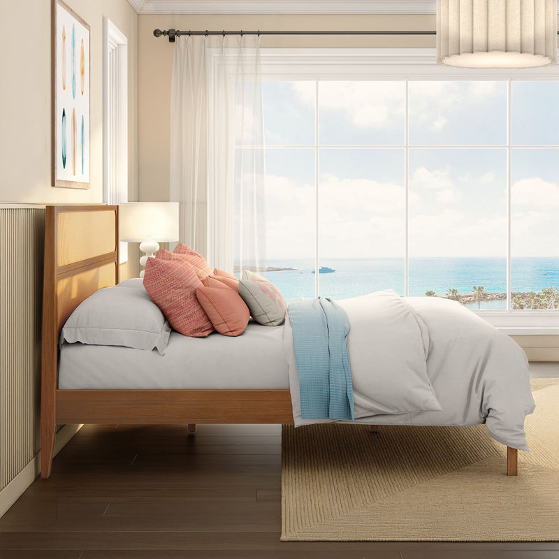 Coastal bedroom interior with wooden rattan headboard, layered neutral bedding and coral accent pillows, sheer curtains and a large window showcasing panoramic ocean and beachfront views