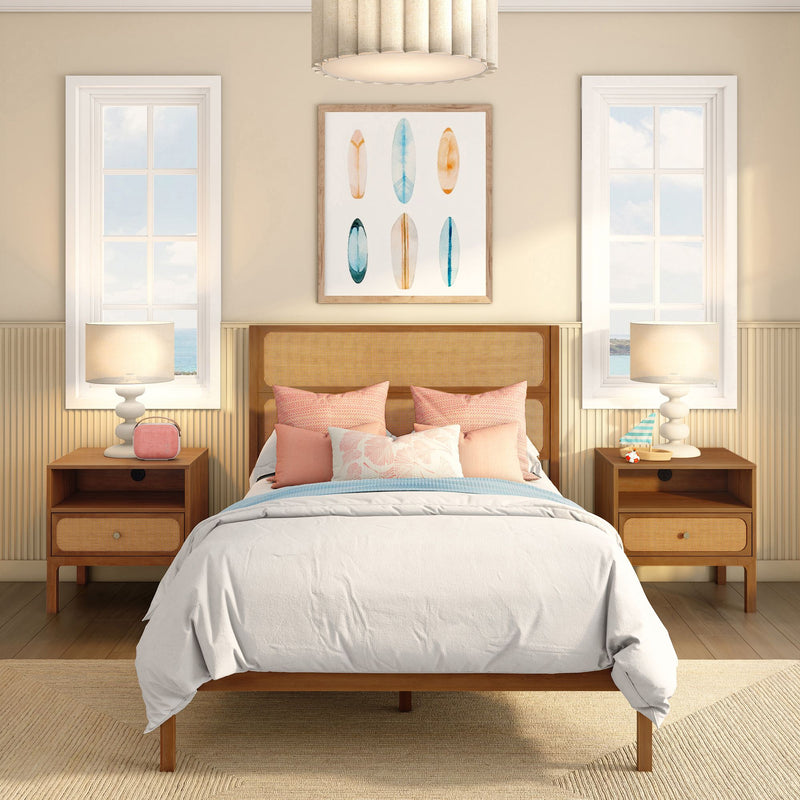 Coastal bedroom decor with rattan headboard, white bedding and pink pillows, wooden nightstands with lamps, ocean-view windows and surfboard wall art