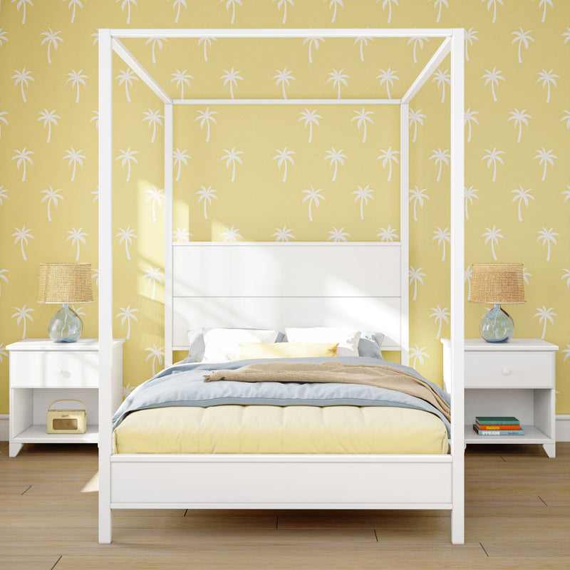 Coastal bedroom interior with white four-poster bed, pastel bedding, yellow palm-print wallpaper, white nightstands and rattan bedside lamps
