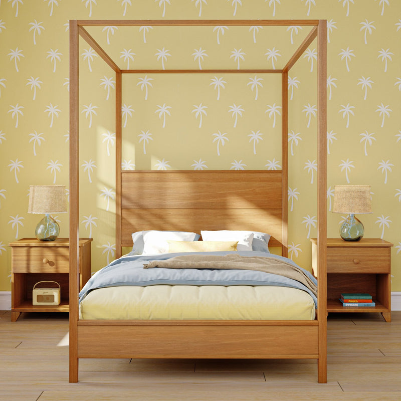 Wooden four-poster canopy bed with pastel bedding in a coastal bedroom featuring yellow palm-print wallpaper, matching nightstands and glass lamps