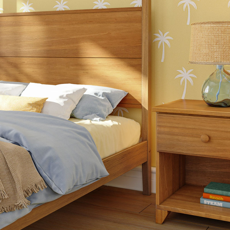 Coastal bedroom close-up with warm wood bed frame and nightstand, blue bedding and neutral pillows, woven lamp and yellow palm-print wallpaper