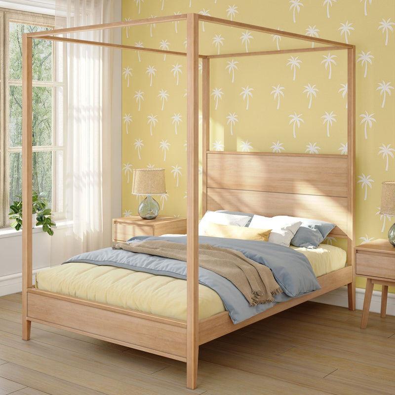 Light wood four-poster canopy bed in coastal bedroom with yellow palm-print wallpaper, rattan lamp, sheer curtains and plant